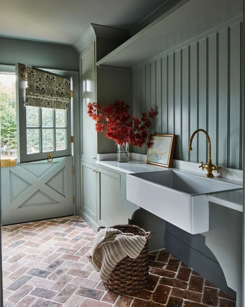 Blue-gray mudroom with pale apron-front sink, patterned window blind, brick-patterned floor and large woven basket.