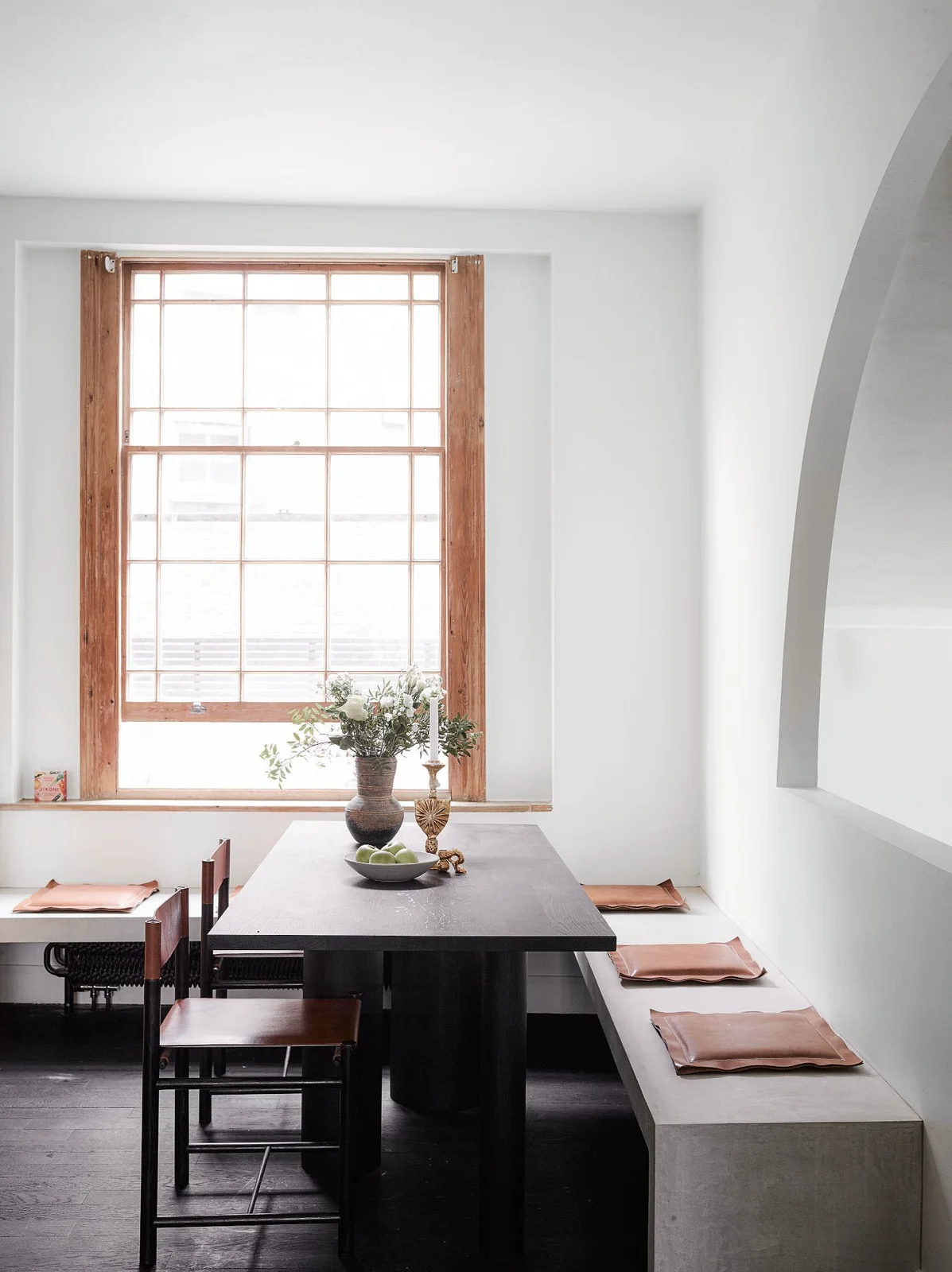 A dark rectangular dining table stands beside a pale built-in bench with small coral cushions and narrow wood chairs beneath a wood-framed window and an arched wall opening