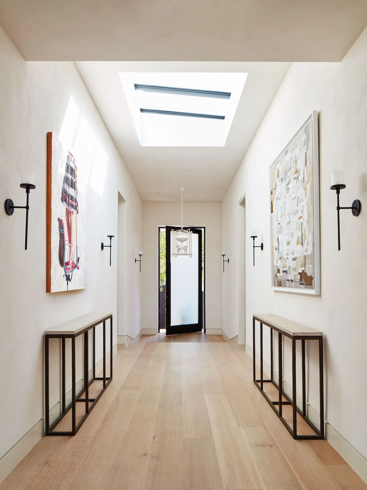 A long pale hallway has black wall-light arms, slim dark-framed consoles, large artwork and a dark door below a broad skylight