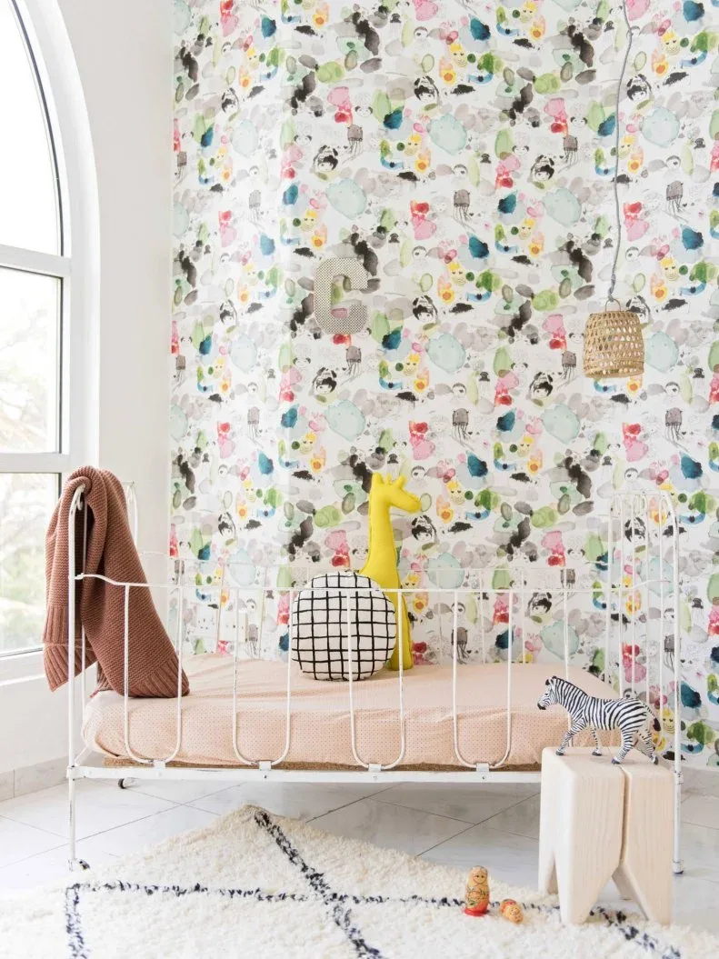 Nursery with lively multicolored wallpaper, pale crib with staged cushions and toys, woven pendant shades and quieter pale rug.