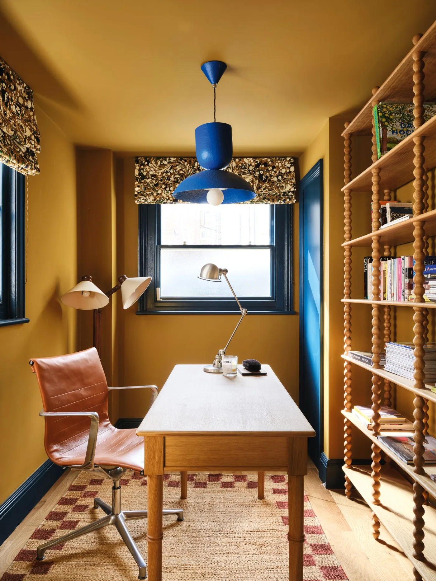 Home office with ochre walls and ceiling, wooden desk and turned shelving, tan rolling chair, blue pendant and dark patterned window shades.