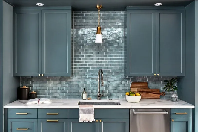 Blue-gray framed kitchen cabinets with glossy blue-gray rectangular tiles rising between upper cupboards above a white sink and counter.