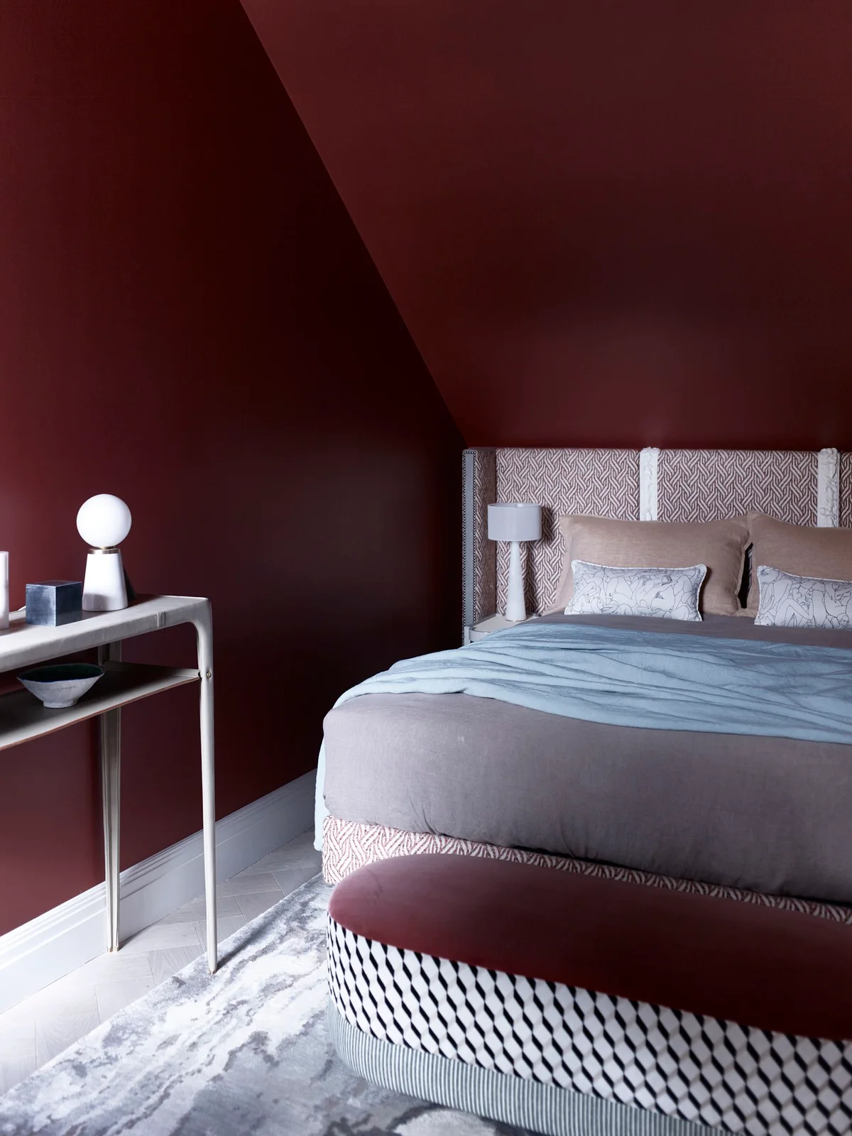Claret walls and a sloping ceiling surround a geometric upholstered headboard, pale bedding and a burgundy bench with a patterned base