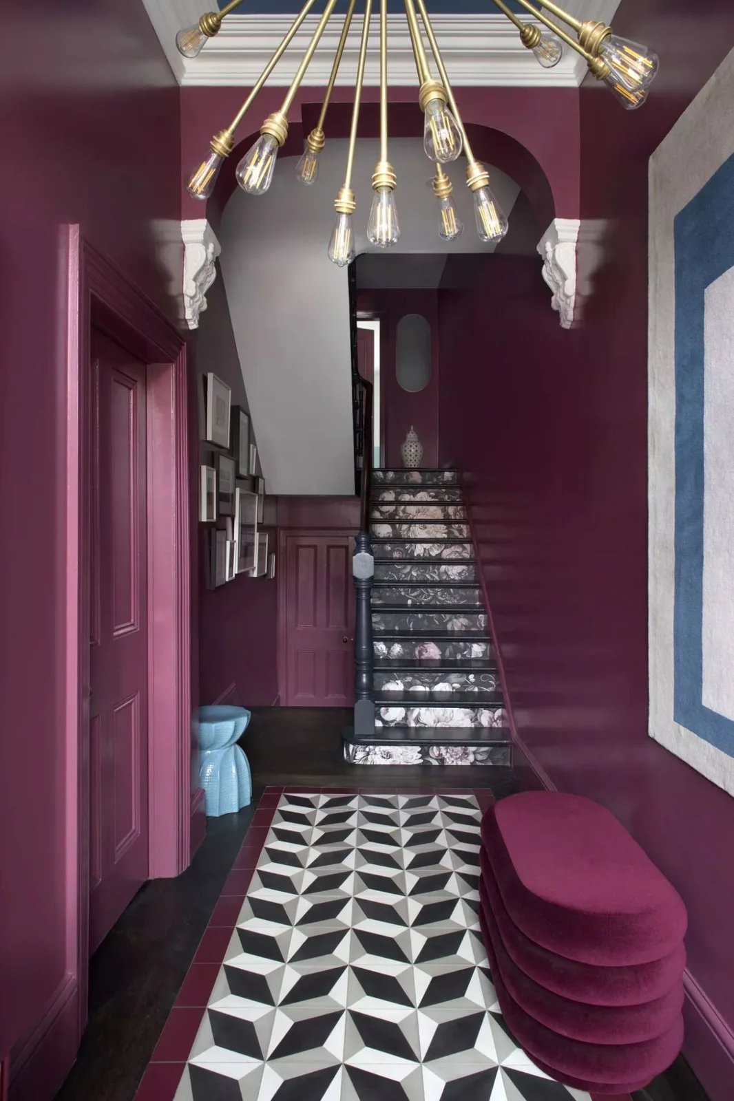 Plum walls surround a black-and-white geometric floor, patterned stair risers, a pink stacked bench and a branching gold-colored light
