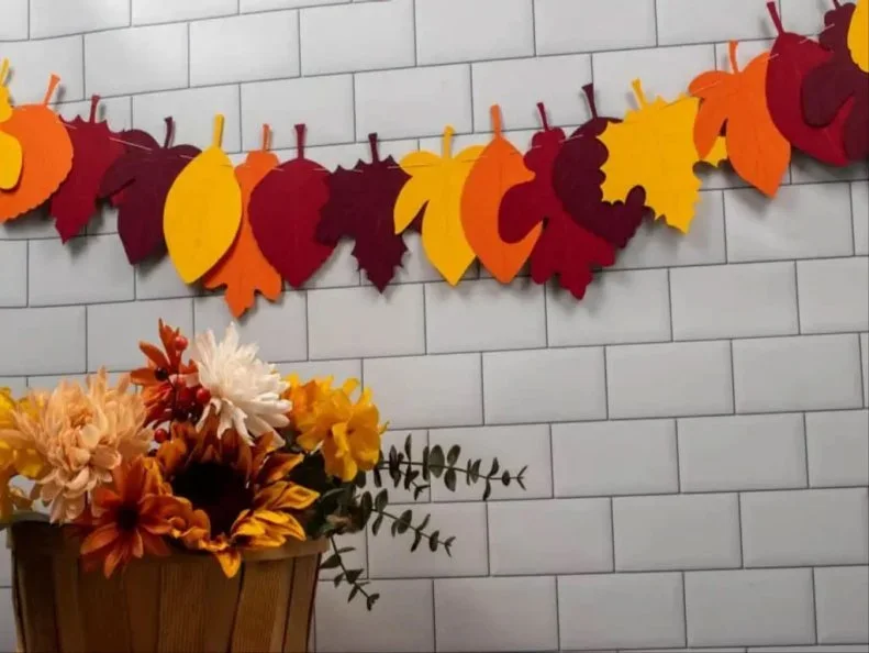 Orange, yellow and deep red paper leaf shapes hang from small clips on a cord against pale rectangular wall tiles