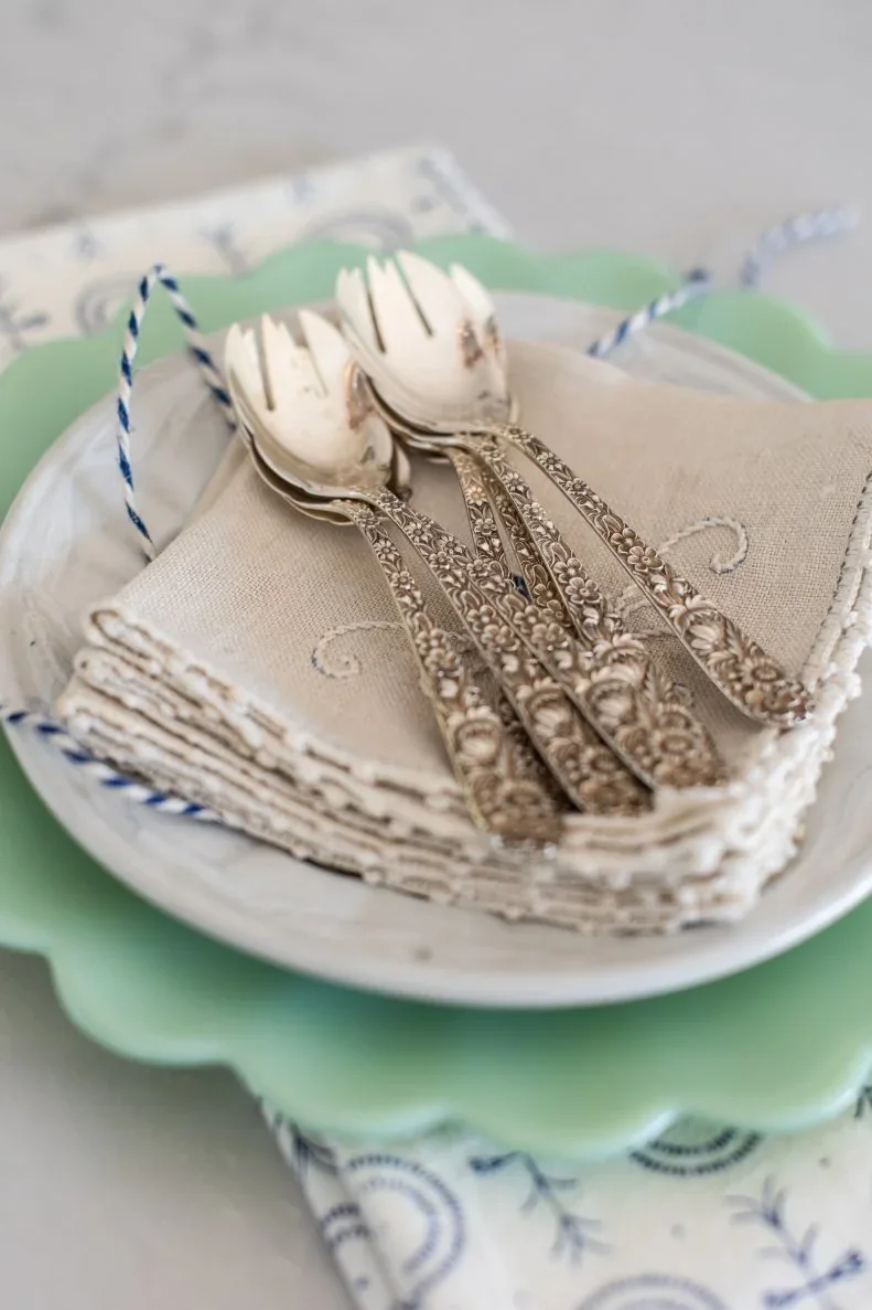 Silver-colored cutlery rests on an embroidered pale napkin above a white plate and scalloped mint-green charger