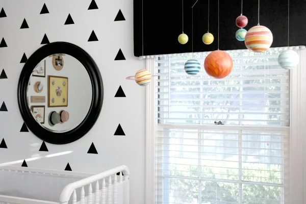 A dark round mirror sits against a pale nursery wall with black triangles beside white window blinds and small hanging colored shapes