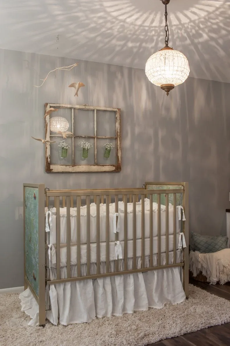 Muted gray nursery with warm open-frame crib, pendant, decorative wall frame, bird-shaped hangings and heavily staged pale bedding.