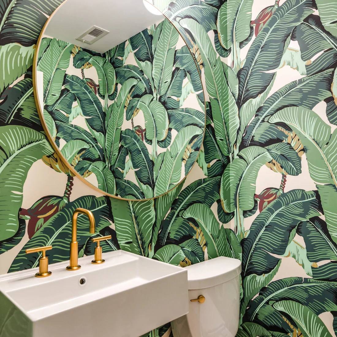 Oversized green leaf wallpaper surrounds a round gold-colored powder-room mirror above a white rectangular sink beside a white toilet