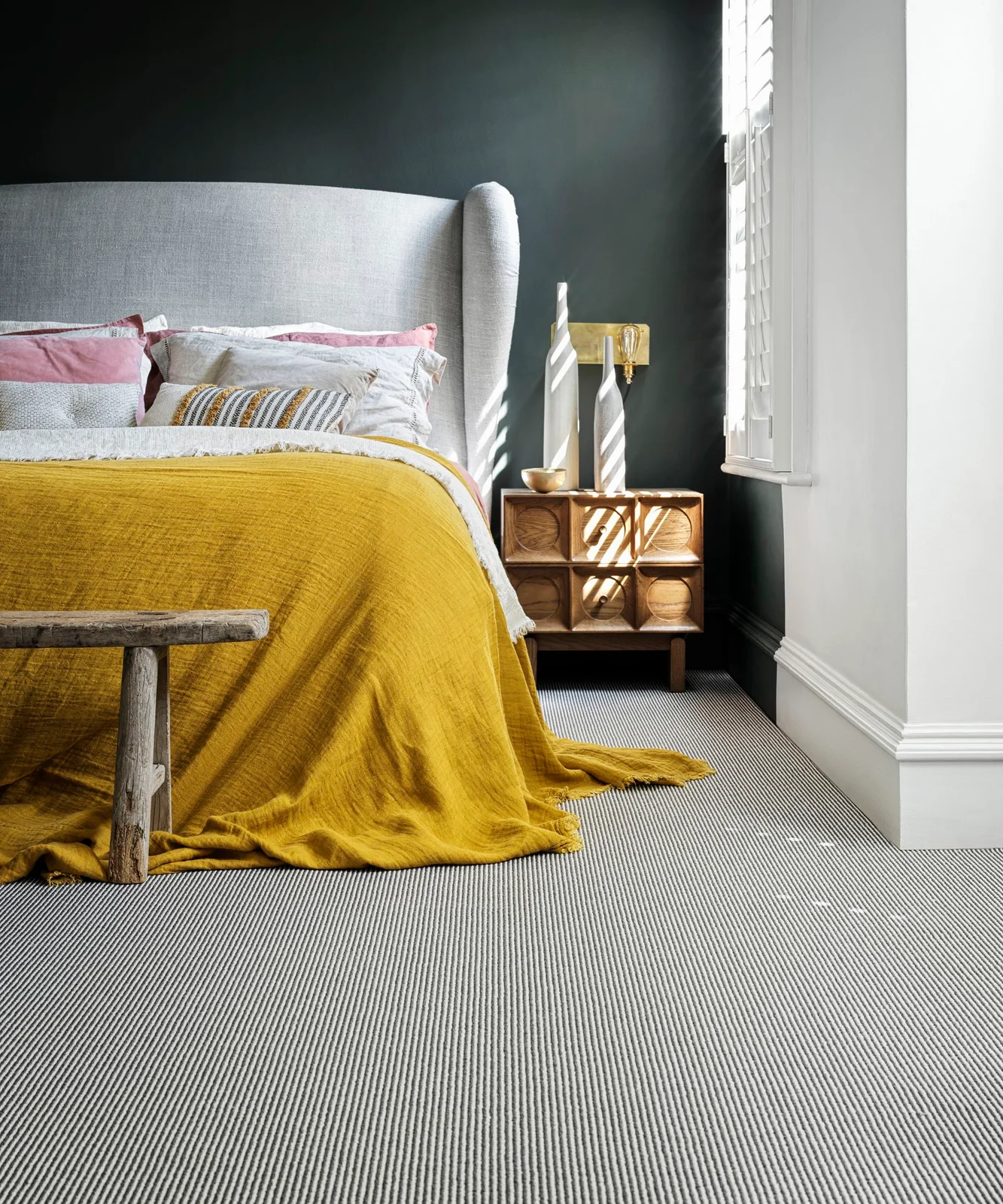 A mustard-yellow bed cover sits below a pale winged headboard beside a dark wall, white shutters and wooden bedside drawers