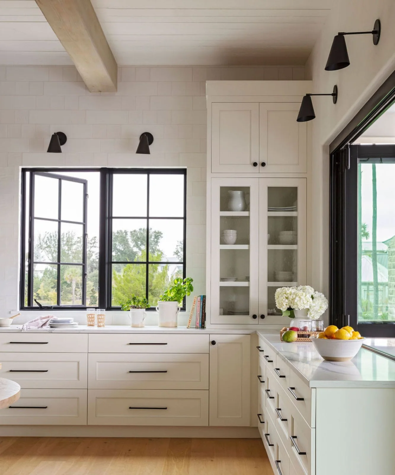White framed cupboards and long drawers with dark handles sit beside glass upper doors, dark-framed windows and simple dark wall lights above pale counters