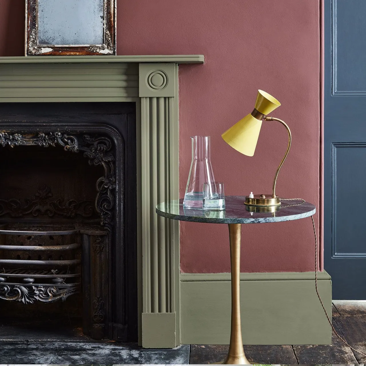 Olive green mantel beside pink wall, small reflective frame and yellow lamp on a round side table