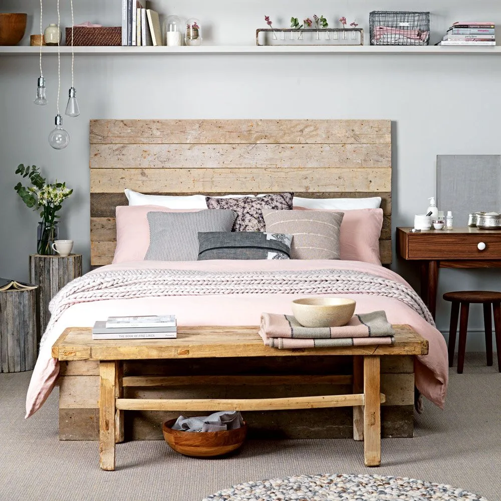 Gray bedroom with a broad wooden headboard, pale pink bedding, wooden bench and a narrow shelf above the bed.