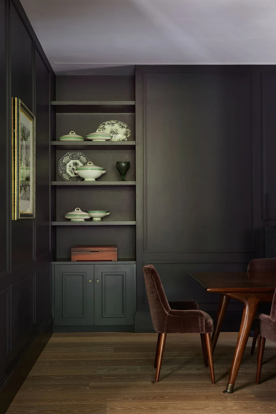 A dark paneled dining alcove holds pale patterned dishes on open shelves above closed cupboards beside a brown upholstered chair and wooden table