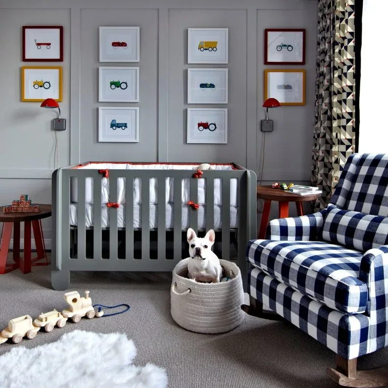 Checked adult chair beside gray crib and paneled wall with vehicle-picture group, red wall lights and simple tables.