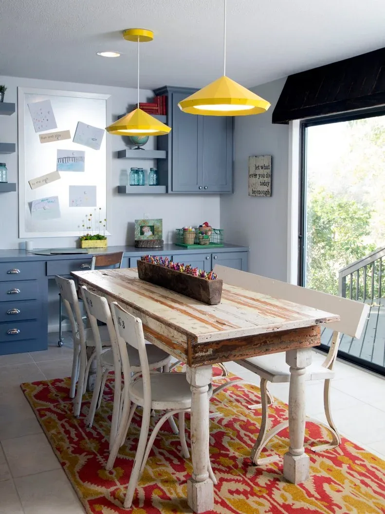 Country craft room with blue cupboards, worn-looking wood table, white chairs, yellow pendants and colorful rug