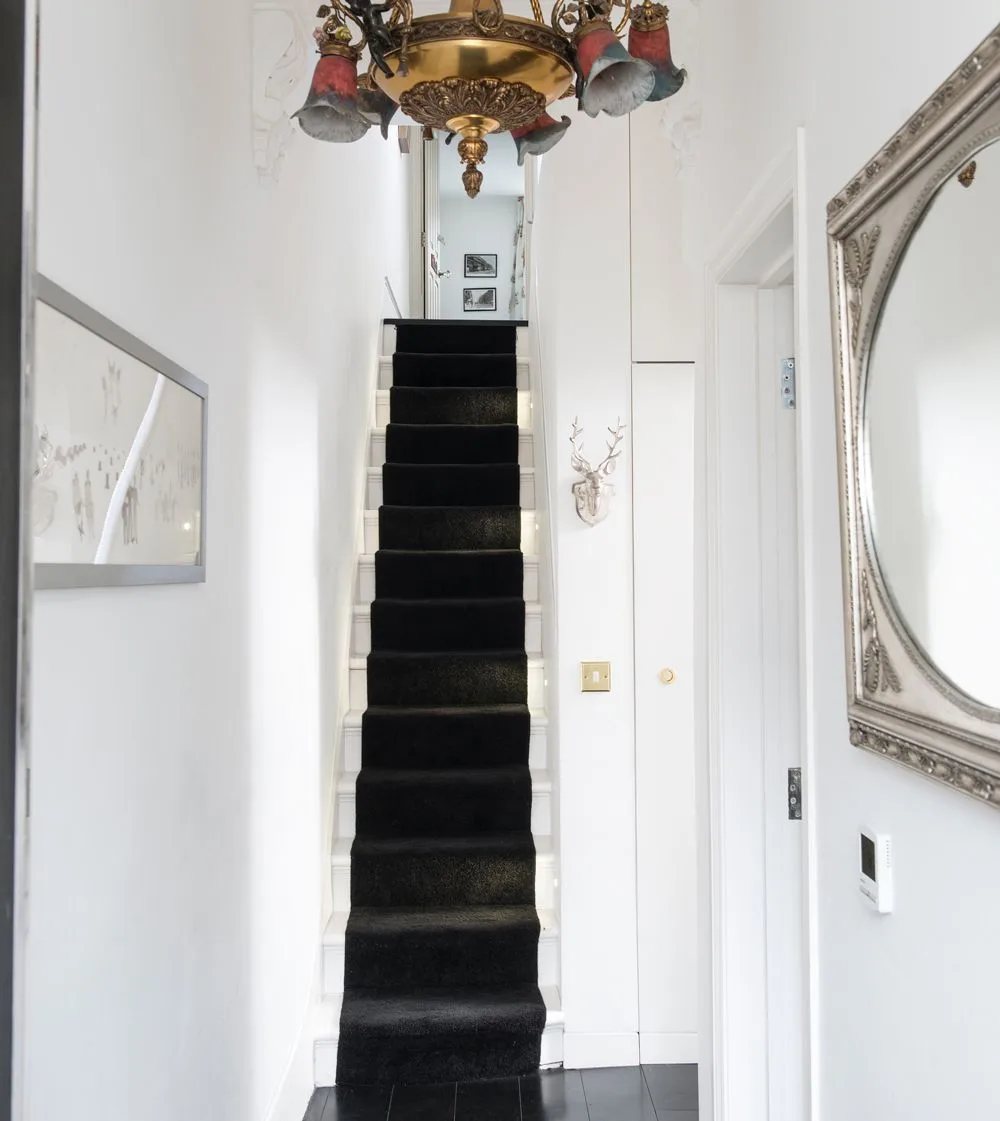 Narrow white hallway with a dark stair runner, ornate ceiling light and decorative framed mirrors on the walls.