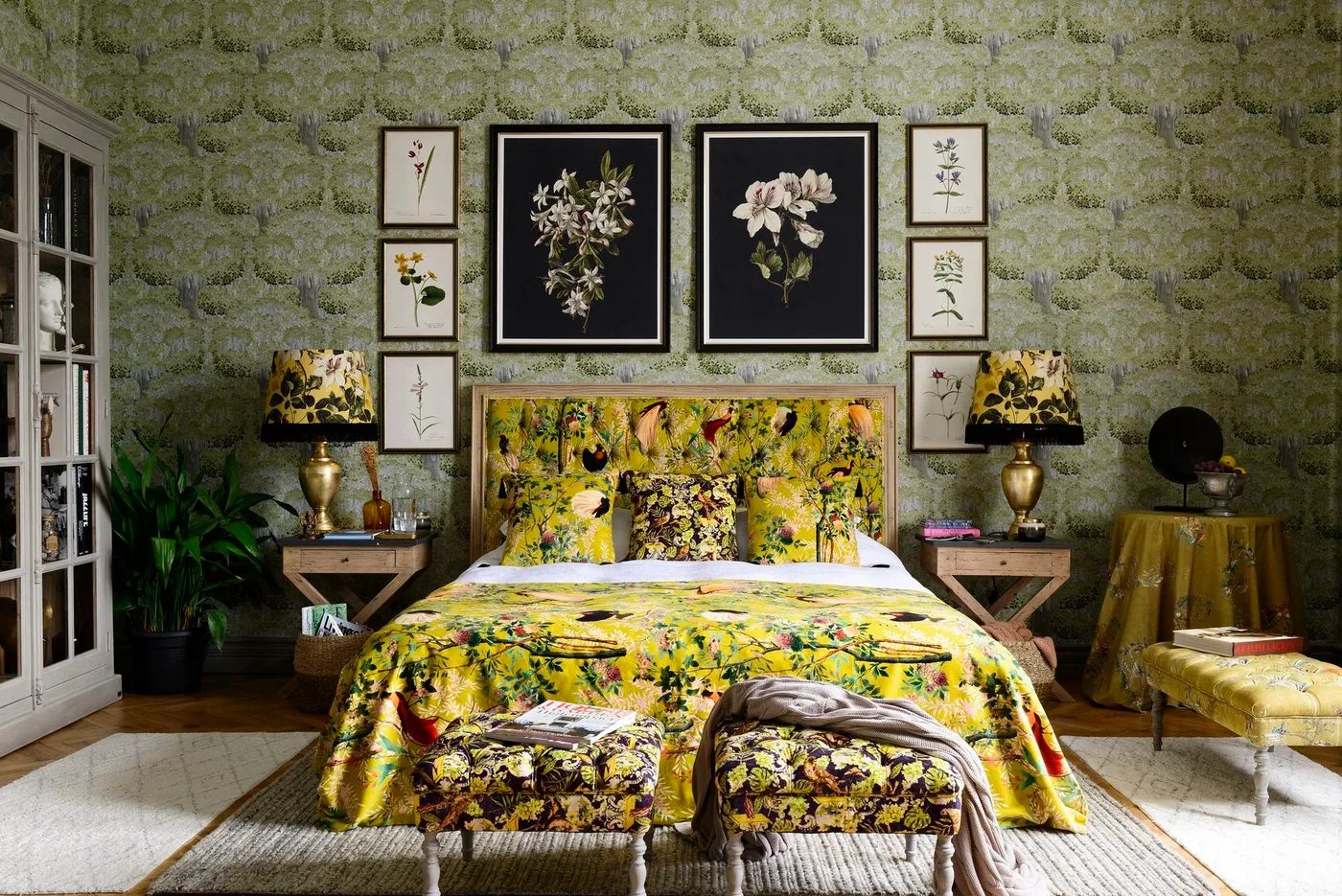 Green tree-pattern bedroom wallpaper behind framed botanical pictures, yellow patterned bedding and matching lamps.