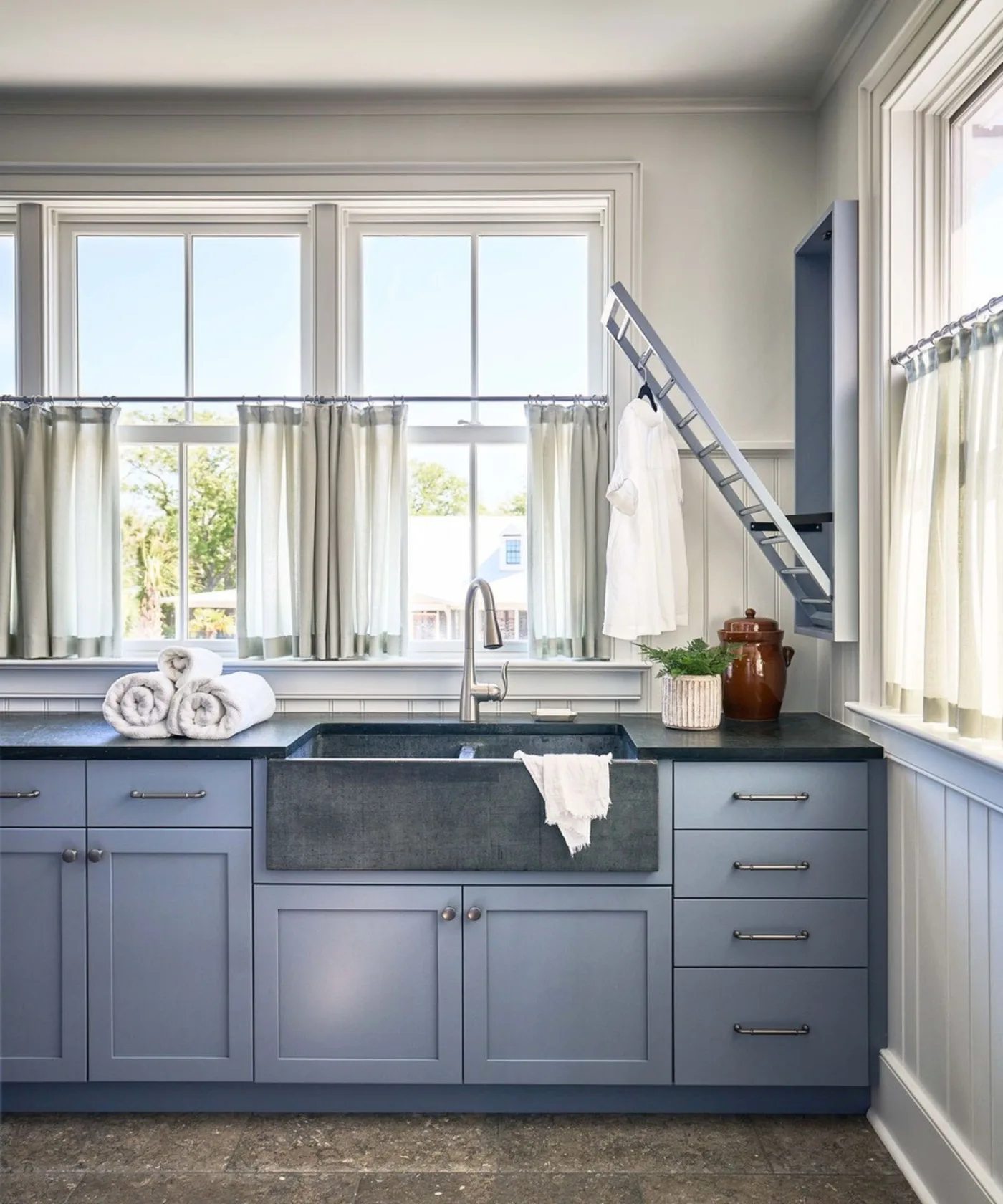 Blue-gray laundry cabinetry with broad dark apron-front sink, pale window curtains, towels and a folding wall rack