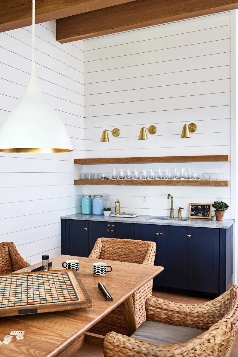 Wood-toned floating shelves over navy cabinets behind a wooden table with woven chairs and a board game