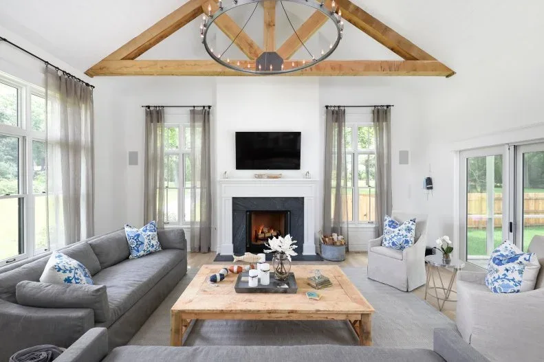 Bright country room with exposed timber truss, dark sofas, pale armchairs, wood table, blue patterned cushions and fireplace television wall