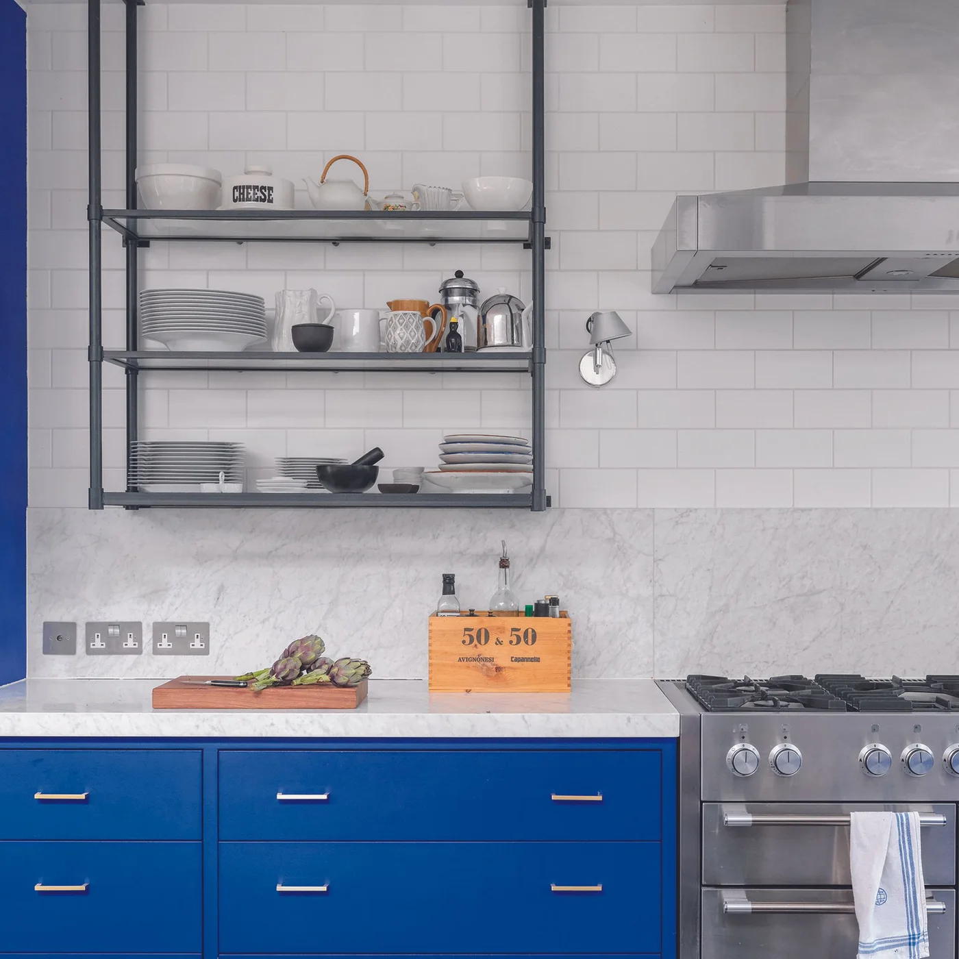 White offset rectangular tiles cover a kitchen wall behind dark open shelves, blue cabinets and a metal cooking hood