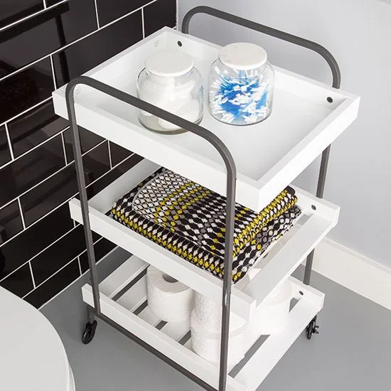 A small white tiered trolley with dark curved rails holds jars, patterned folded towels and toilet paper beside glossy dark wall tiles