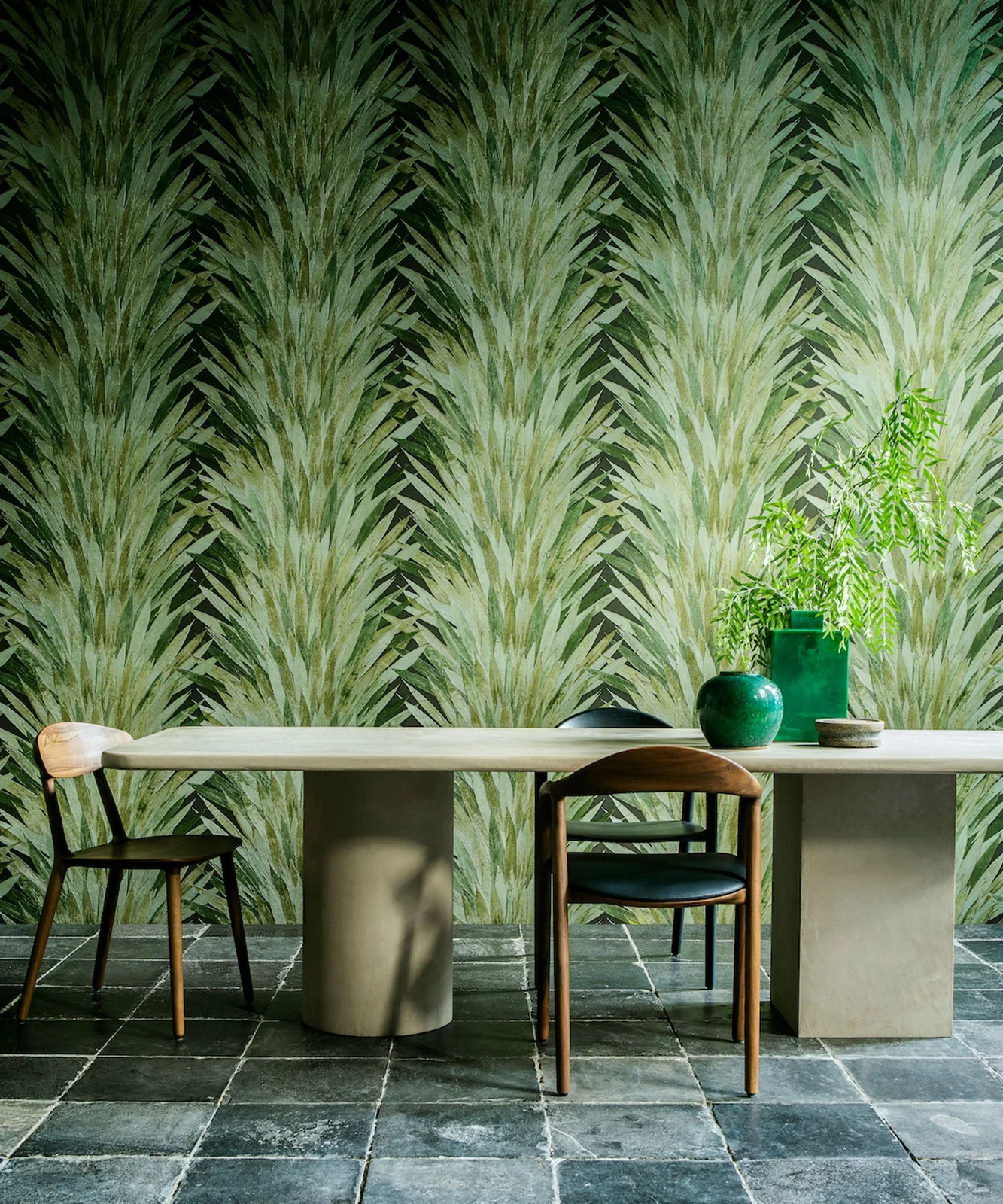 Large green leaf-patterned wallpaper stands behind a pale table with wooden chairs, dark seat pads and green vessels