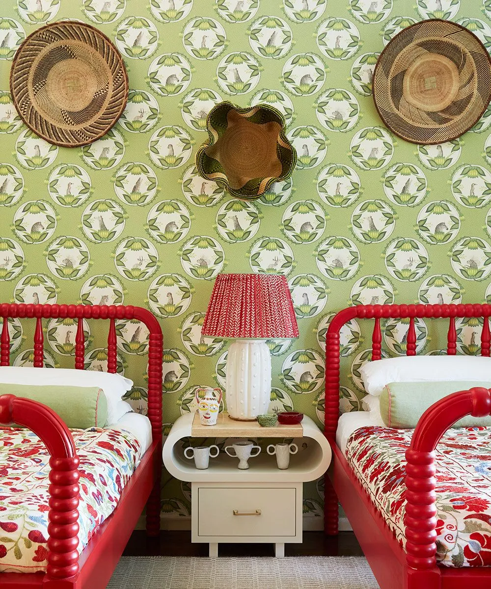 Two red turned bed frames with floral bedding, green botanical wallpaper, woven hats, small mirror and pale central bedside cabinet.
