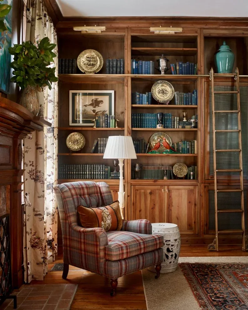 Plaid upholstered chair beside wood bookcases with collected plates, a library ladder, floor lamp and patterned curtains