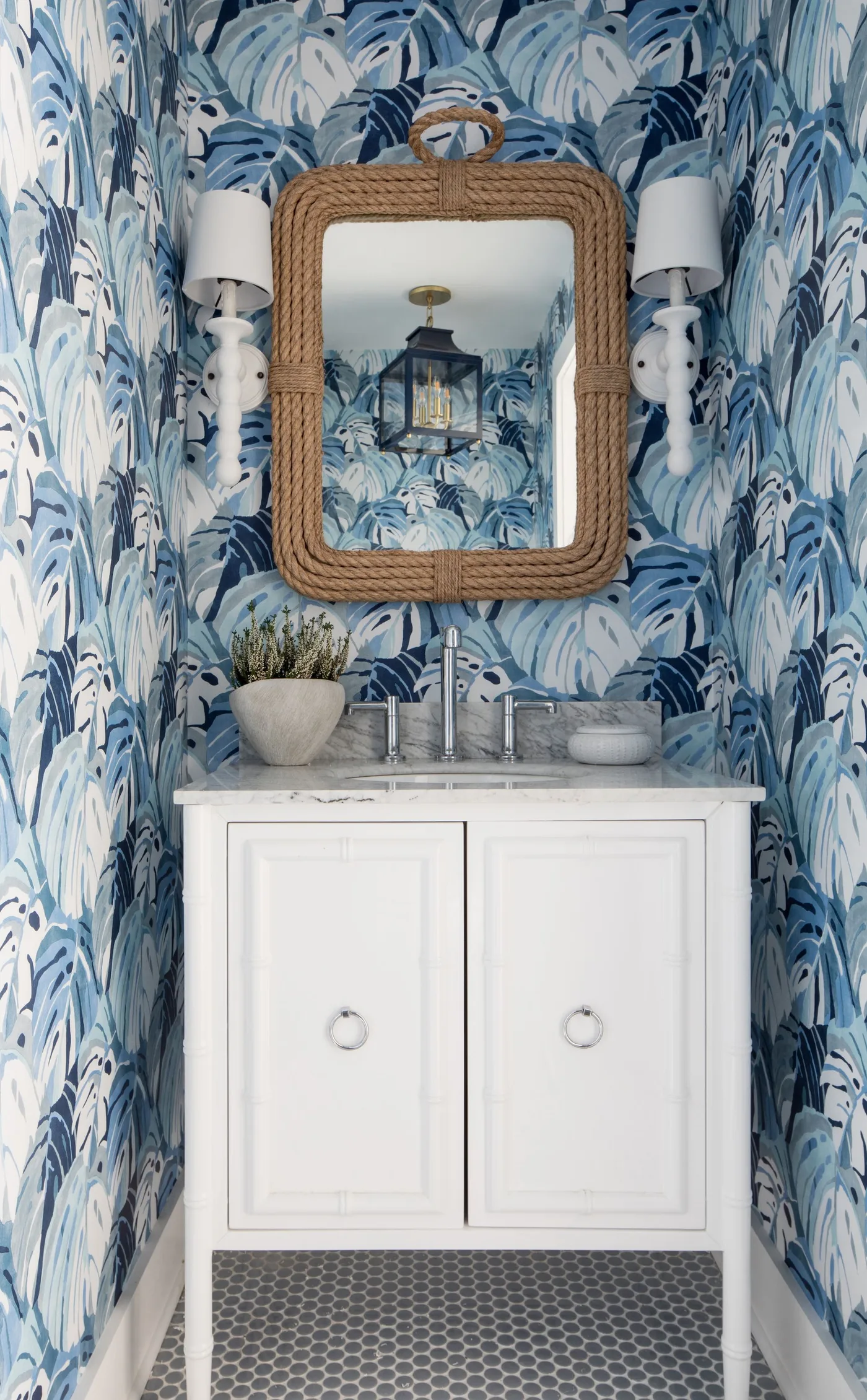 Powder room with blue leafy wallpaper, rectangular woven mirror, white vanity, paired white-shaded lights and small plant beside the basin.