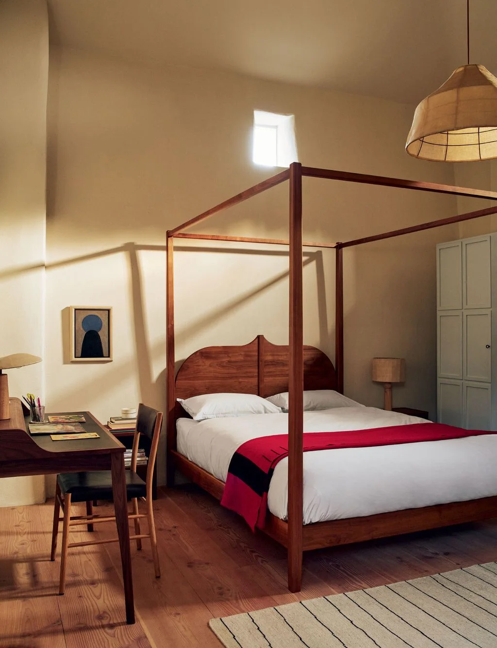 An open wood four-poster bed with pale bedding and a red throw stands beside a wood desk and chair below pale walls and a shaded pendant