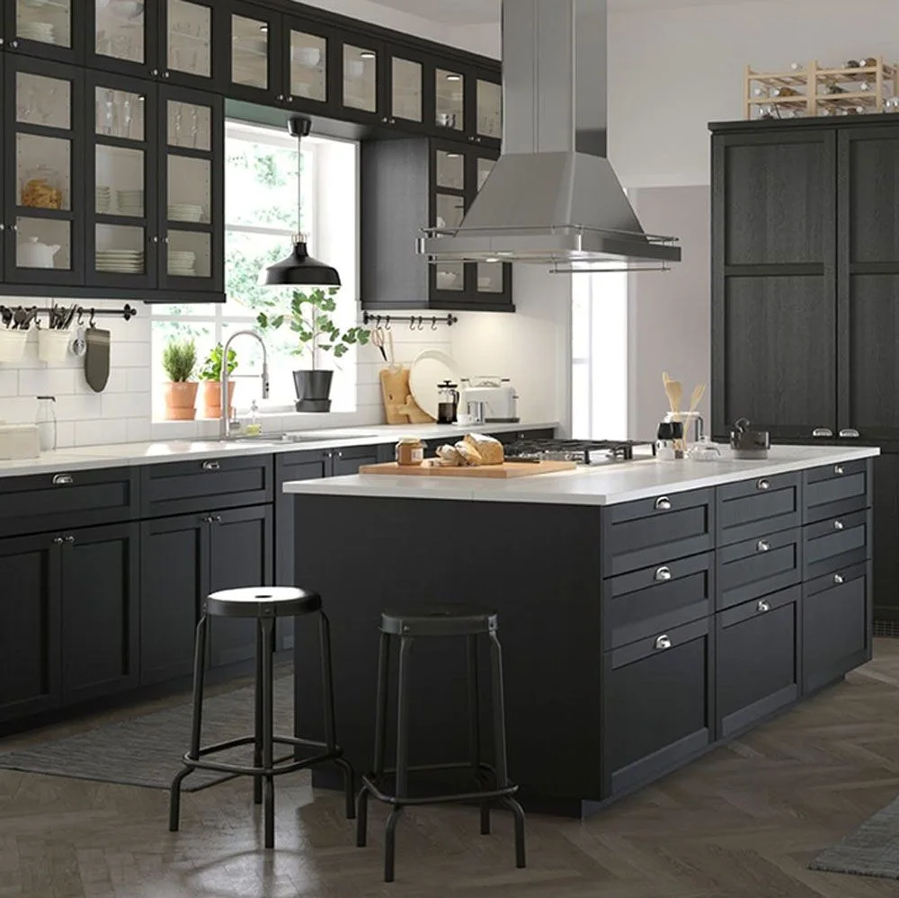 A dark kitchen has glass-front upper cupboards and lower drawers beside a white counter and island top beneath a broad suspended cooker hood