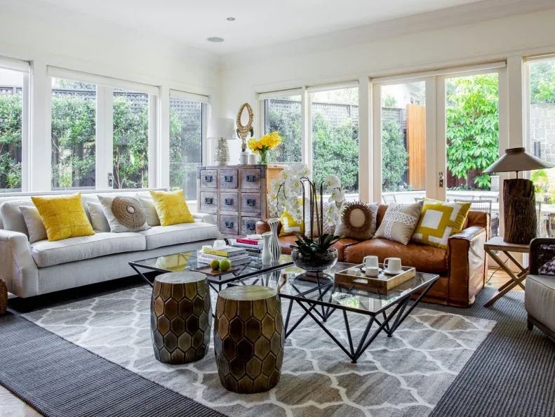 Bright living room with pale sofa, tan armchairs, two glass-topped tables with dark geometric bases, yellow cushions, small golden stools and many windows.