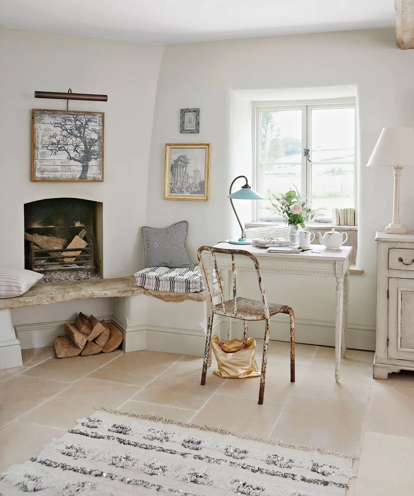 Small arched fireplace opening with pale hearth bench, cushions, lower logs and a desk beside a bright window