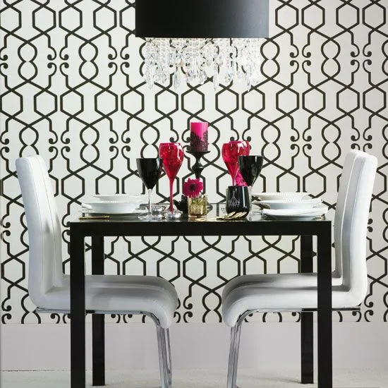 A black dining table and white chairs stand against black-and-white geometric wallpaper beneath a dark pendant with hanging clear details and red glassware