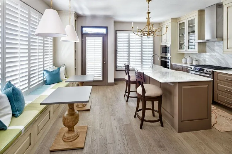 A paneled island with upholstered stools stands beside a long window bench, pale shaded pendant lights, framed kitchen cupboards and a small pedestal table