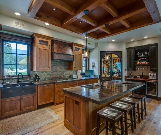 A timber island and cabinet wall sit beneath an exposed wood ceiling with dark countertops.