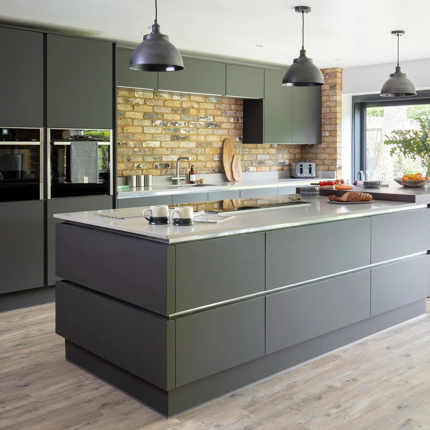 Deep gray-green cabinets and an island stand beside an exposed brick wall, pale counters, built-in ovens and dark dome-shaped pendants