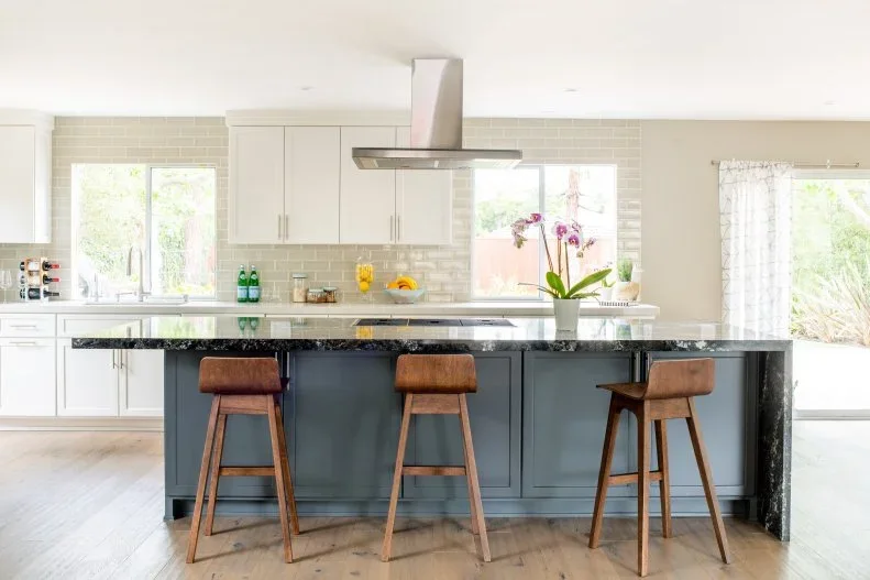 A muted blue paneled island with a dark counter and wood stools faces white cupboards beneath a plain metal extractor hood beside a light rectangular tile wall