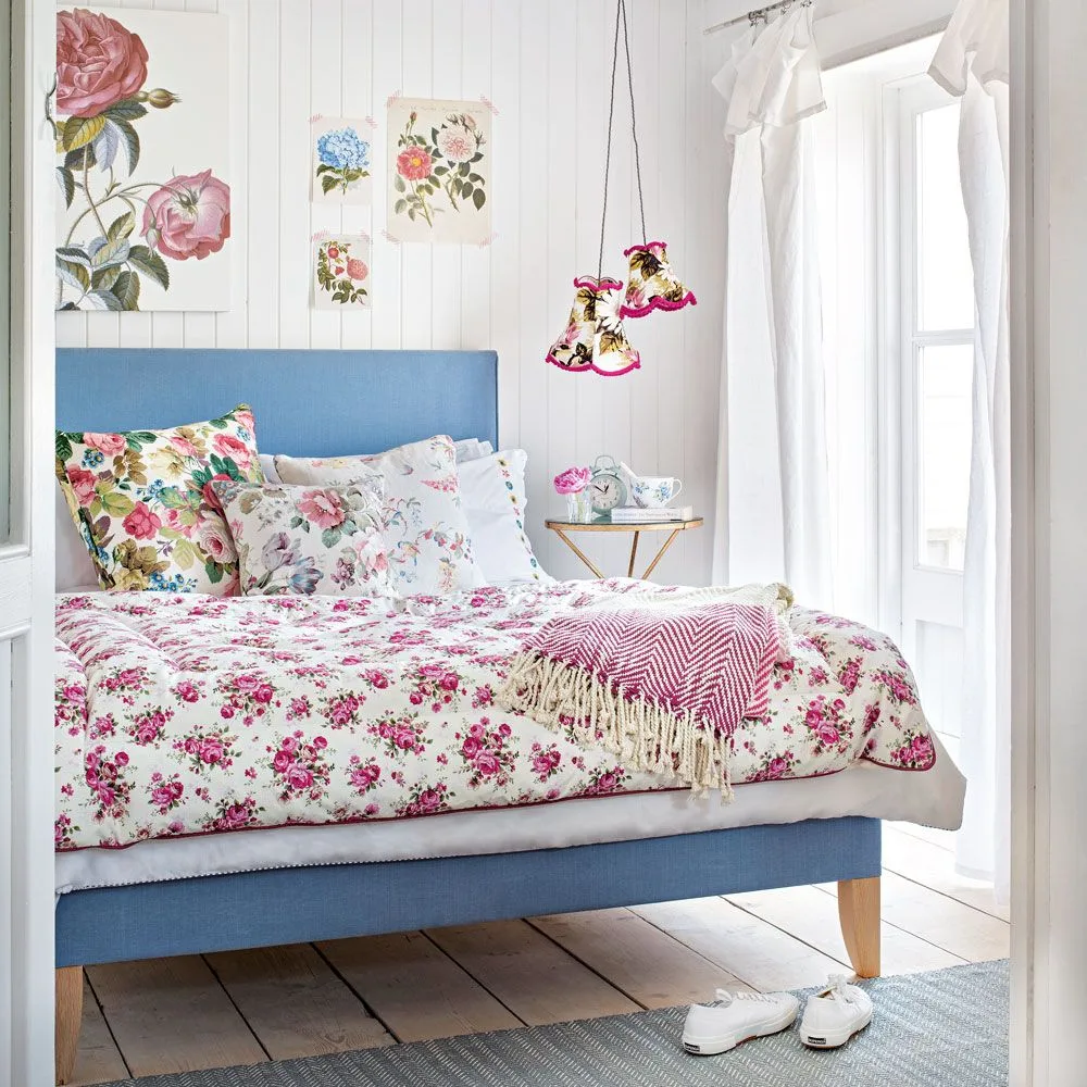 Bedroom with a blue bed, floral bedding and cushions, white vertical wall boards, botanical pictures and small hanging floral shades.