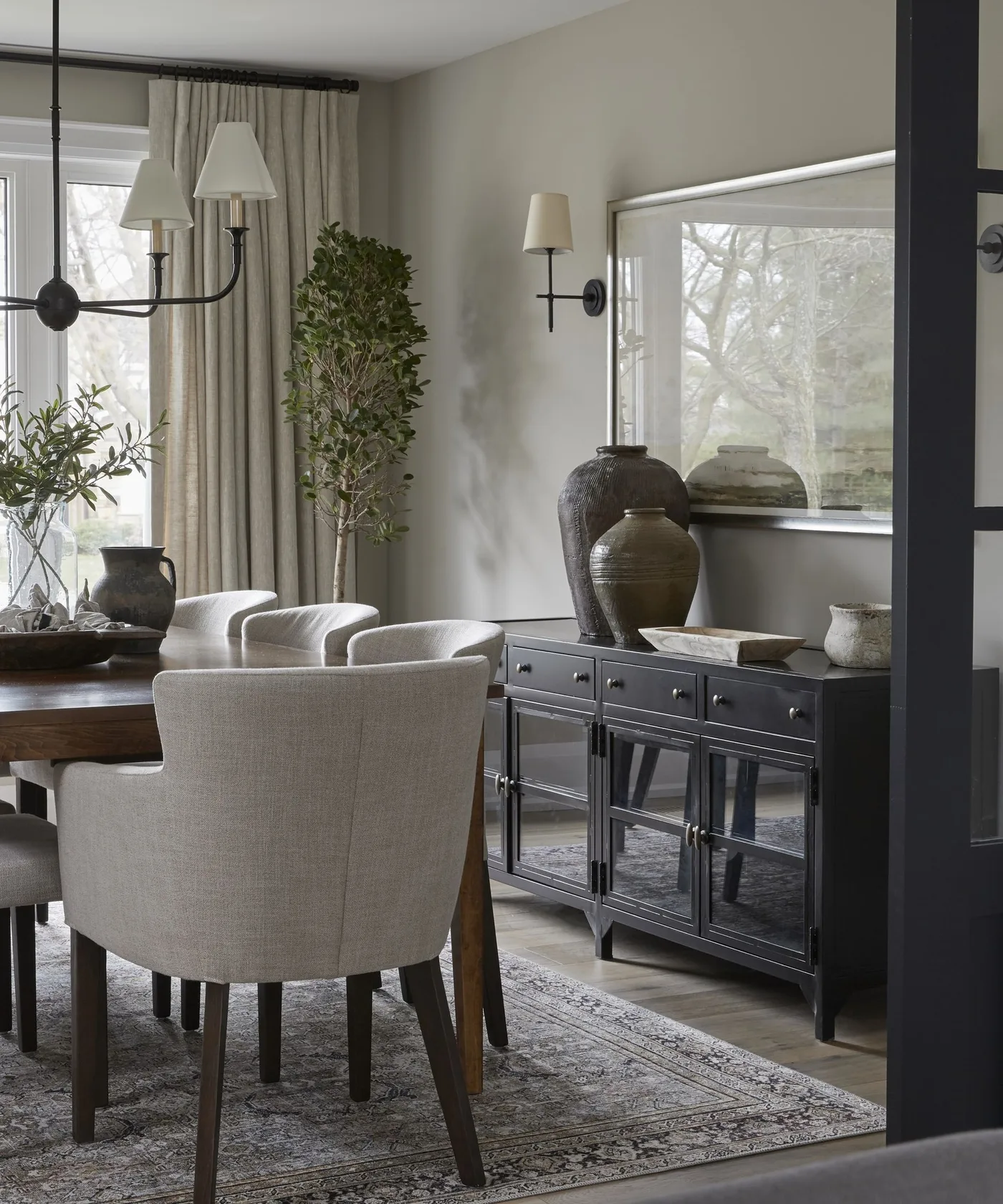 Dark glazed dining sideboard beneath wide artwork beside a wood table, neutral chairs and green plants