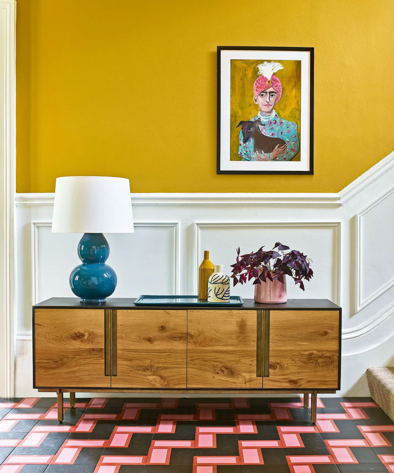 A straight wood sideboard with a dark top and long gold-colored handles stands beside yellow walls, white paneling and a pink-and-dark patterned floor