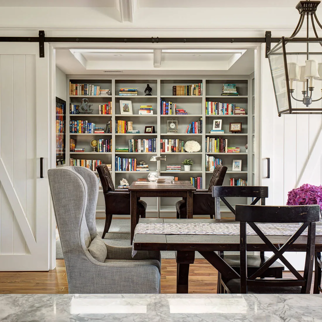 Dining table with cross-back chairs beside white sliding doors opening to a book-lined room and another table