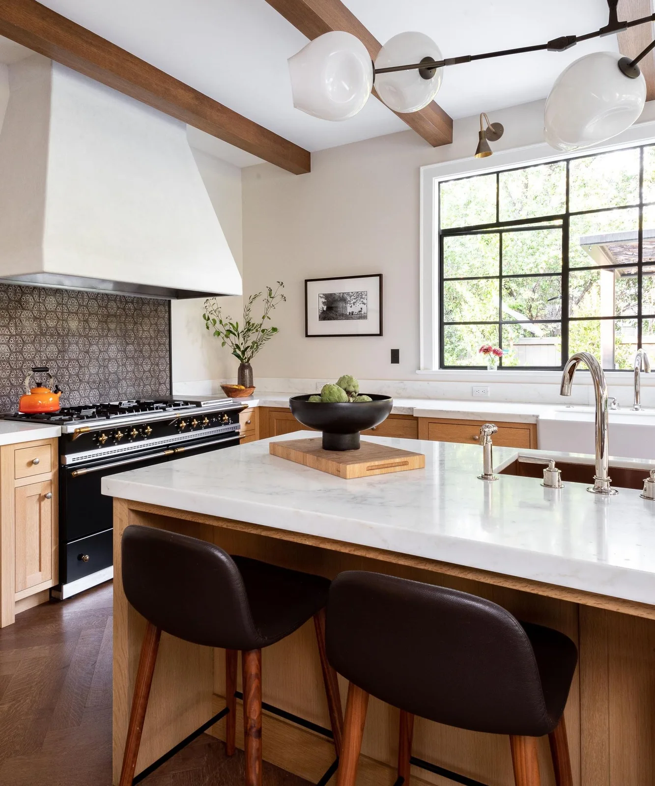 Wood framed cabinets and a broad pale island sit below a plain hood and globe light beside a patterned cooker backsplash, warm ceiling beams and a dark-framed window