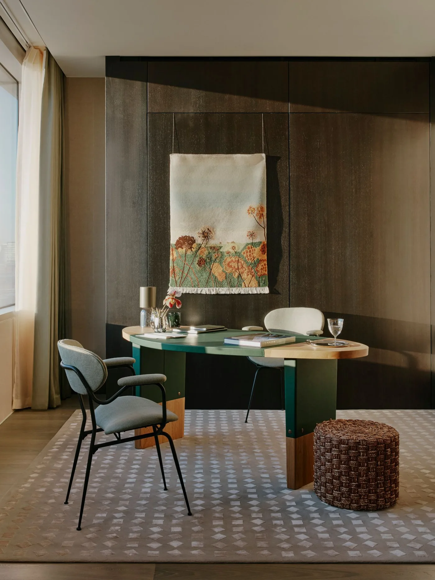 Home office with dark wooden wall panels, oval desk with green and pale surfaces, gray chairs, floral textile wall hanging and woven brown pouf.