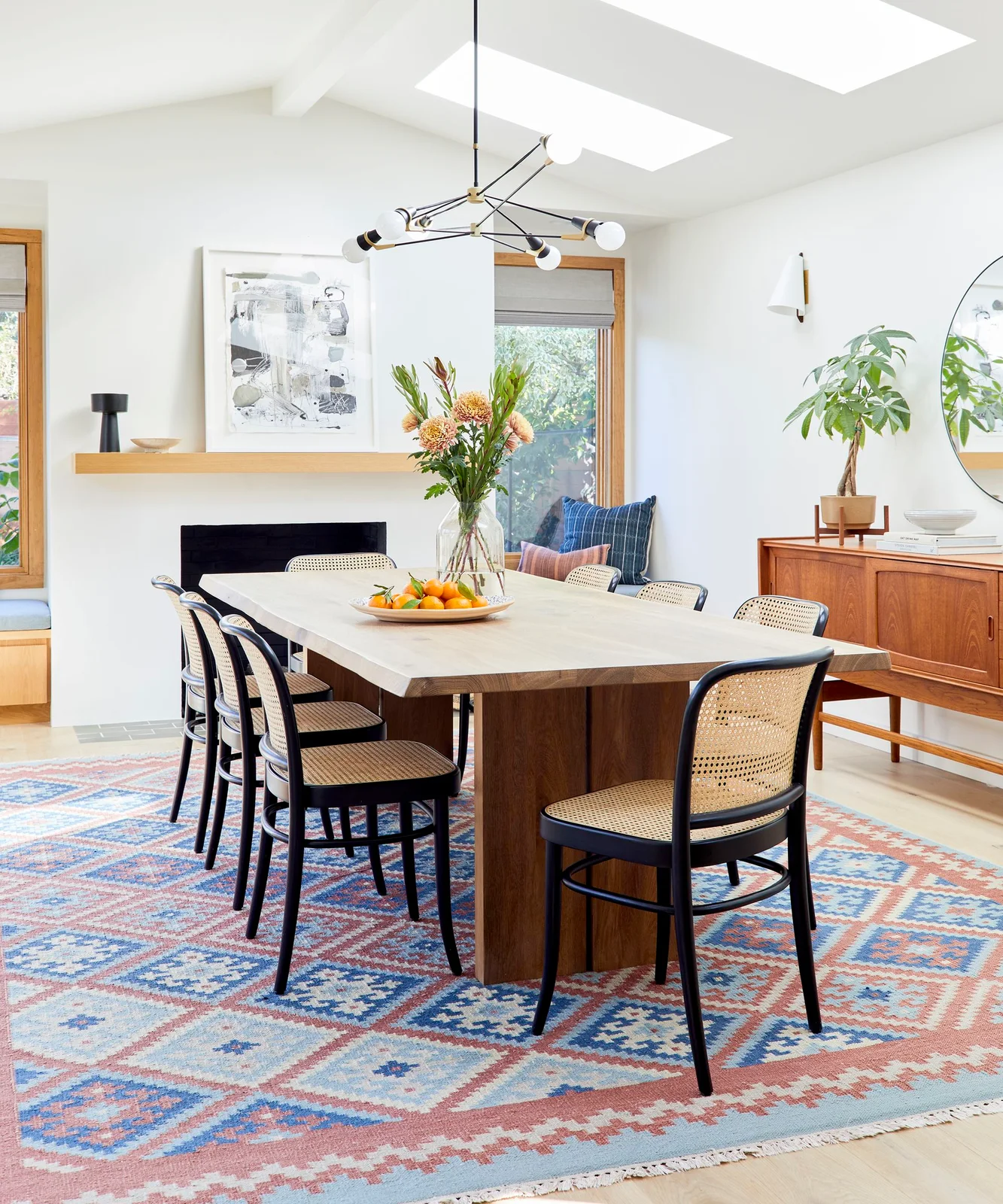 Black-framed woven-back chairs surround a pale table above a blue-and-red patterned rug beneath a branching light and roof windows