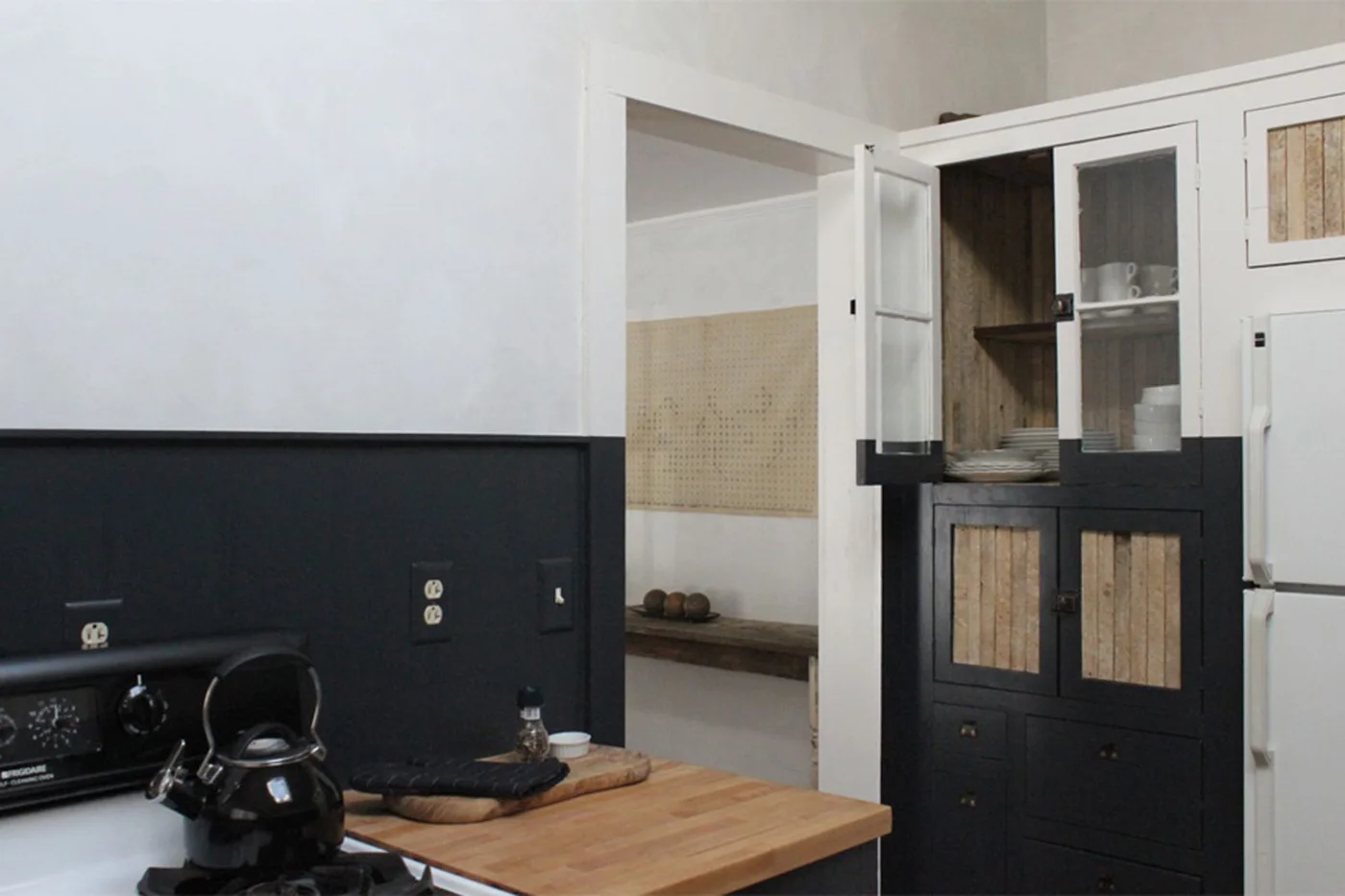 A kitchen has a dark lower wall beside a two-tone glazed cabinet with white upper doors and dark lower doors containing warmer wood panels