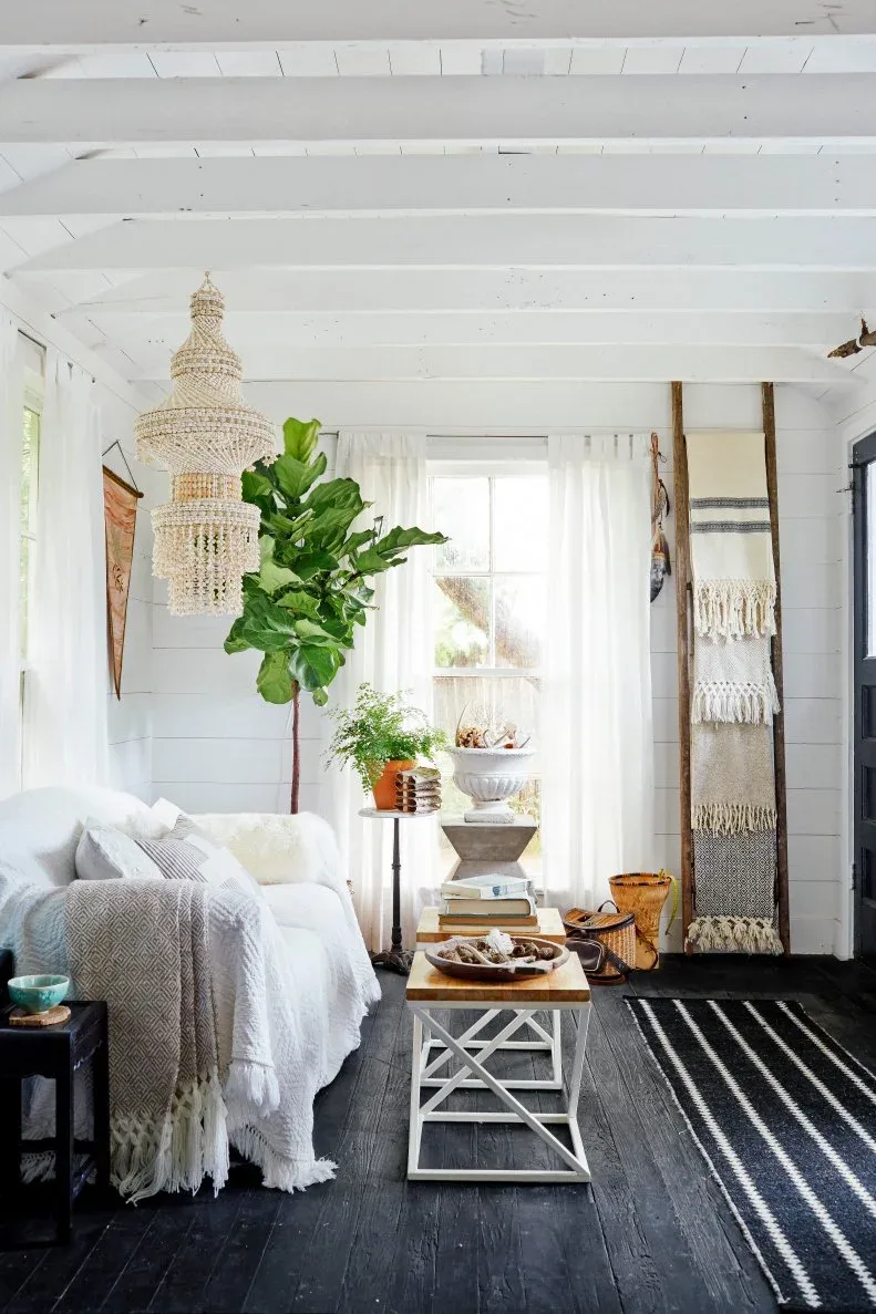 White living room with a tiered chandelier, striped rug, leafy plant and blankets on a leaning ladder.