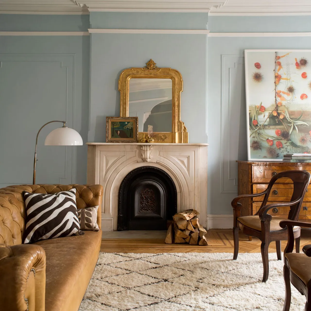 Pale stone fireplace beneath a gold-framed mirror beside a leather sofa and timber armchair